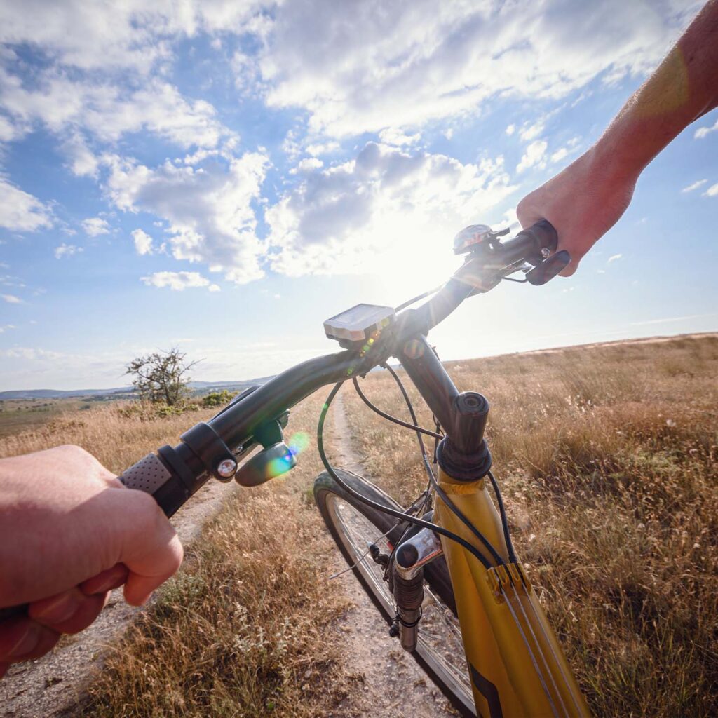 Rider’s point-of-view holding bicycle handlebars while trail riding on a sunny off-road path. - eMotoX Sports