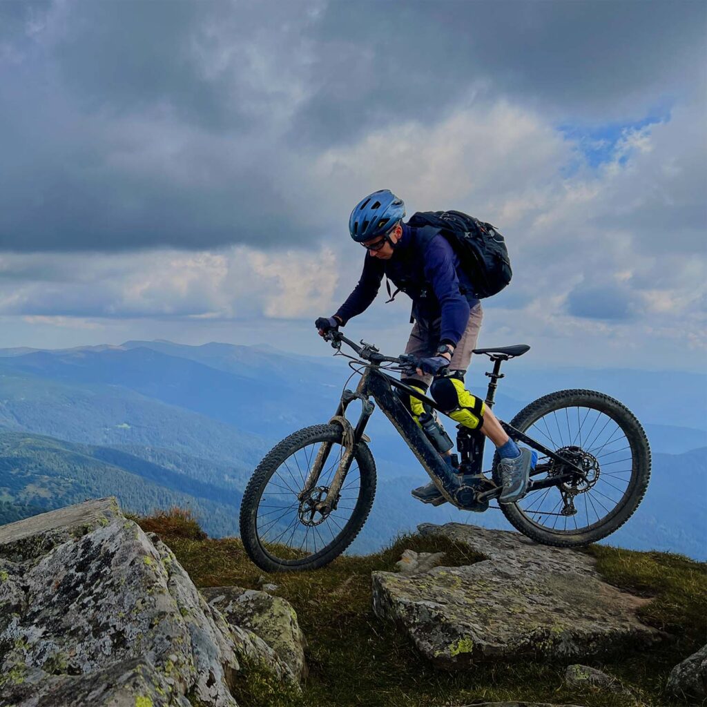Rider on an electric mountain bike navigating a rocky alpine trail with mountain scenery in the background. | eMotoX Sports